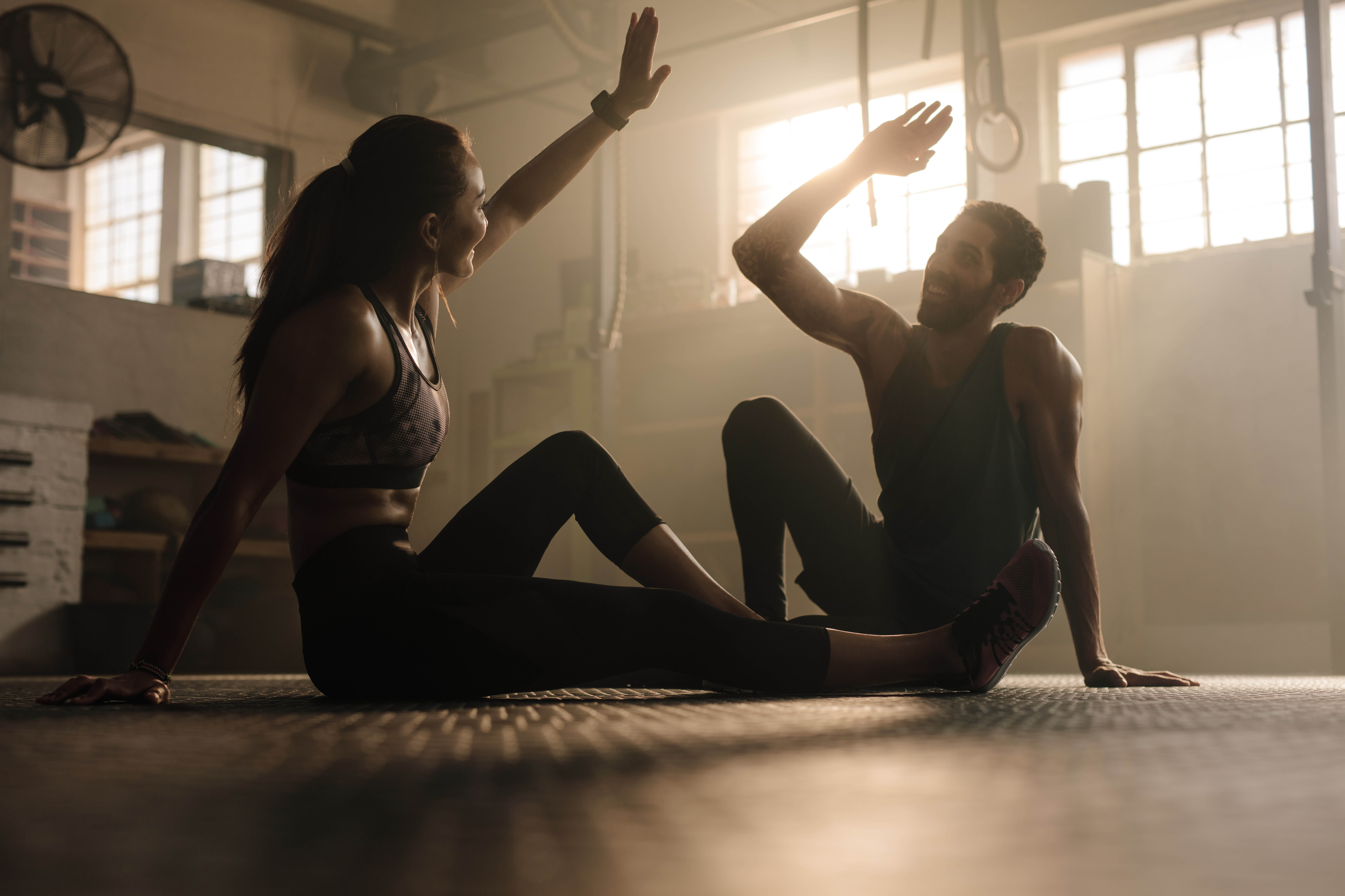 Young, fit couple sitting on the floor of a gym, hands raised as if they're about to high five Young, fit couple sitting on the floor of a gym, hands raised as if they're about to high five